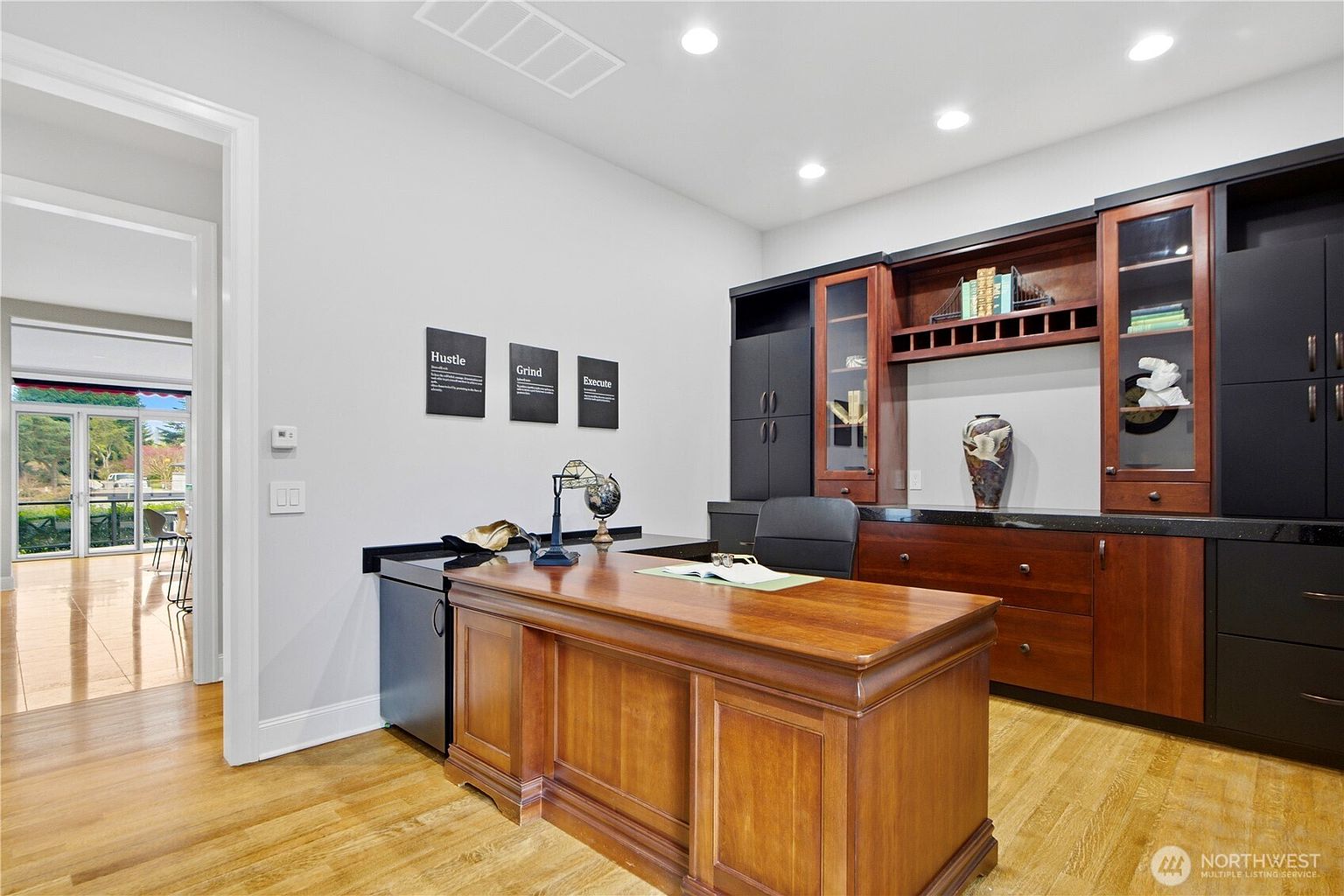 This home office features a large, traditional wood desk positioned centrally in the room, facing a built-in cabinetry unit with dark and wood-toned finishes. The space is illuminated by recessed lighting and includes three framed motivational prints on the wall, while a doorway leads to an adjacent room with a view of an outdoor patio. The room has light-colored walls and wood flooring, creating a professional and organized atmosphere.