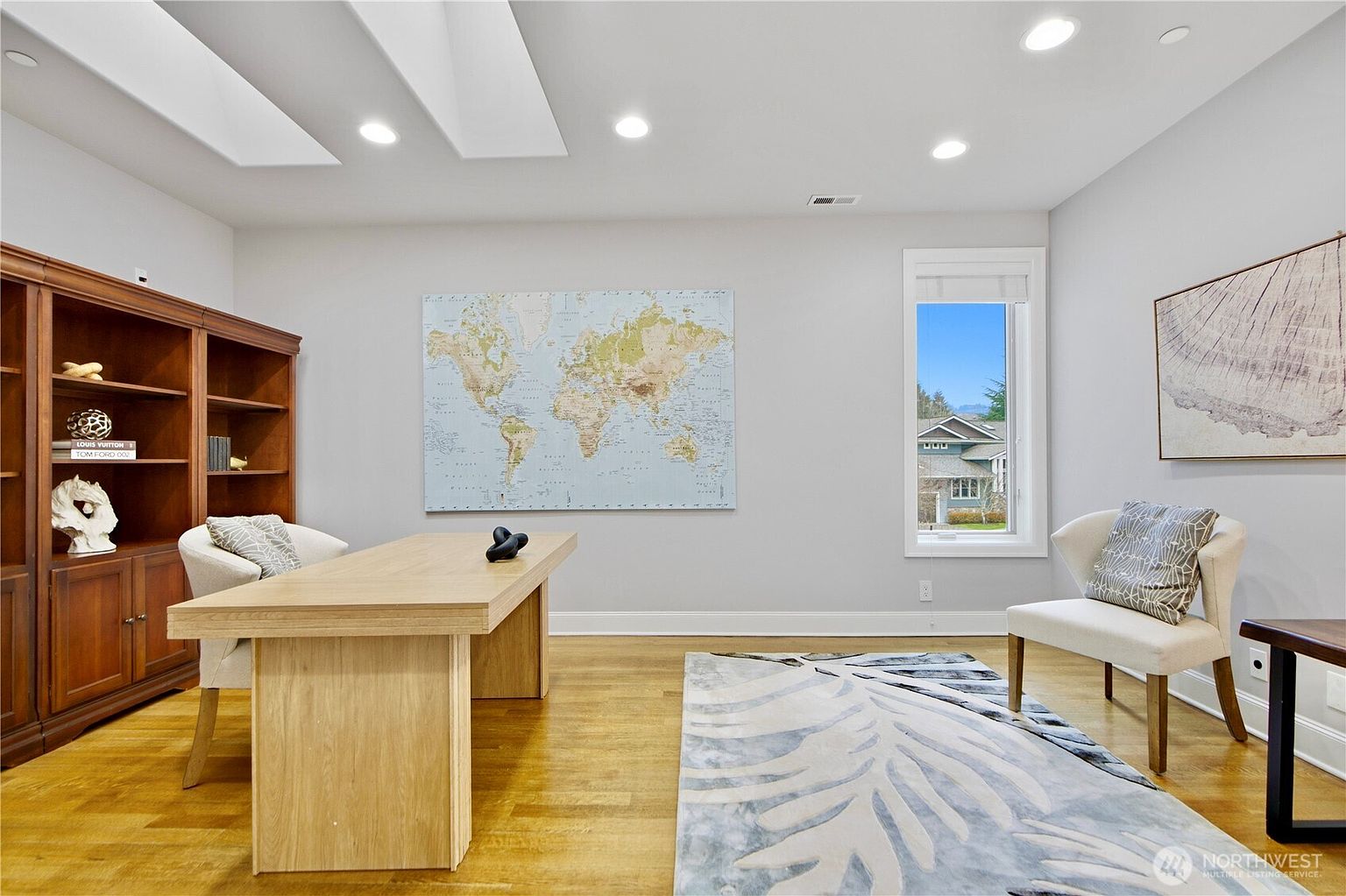 This bright and airy home office features light wood flooring, a large central desk, and a built-in wooden bookshelf along the left wall. A prominent world map serves as a focal point on the main wall, while a comfortable armchair sits near a window that provides natural light and a view of the neighborhood. The room is finished with a patterned area rug and recessed ceiling lighting, creating a professional yet inviting workspace.