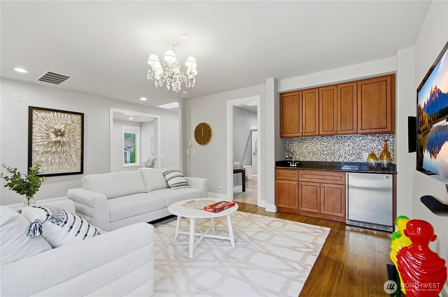 This bright and inviting living room features a neutral color palette with light-toned sofas and a patterned area rug over hardwood flooring. To the right, a built-in wet bar with wood cabinetry, a dark countertop, and a mosaic tile backsplash adds functionality to the space. The room is illuminated by a decorative chandelier and includes a wall-mounted television, creating a comfortable area for relaxation and entertainment.