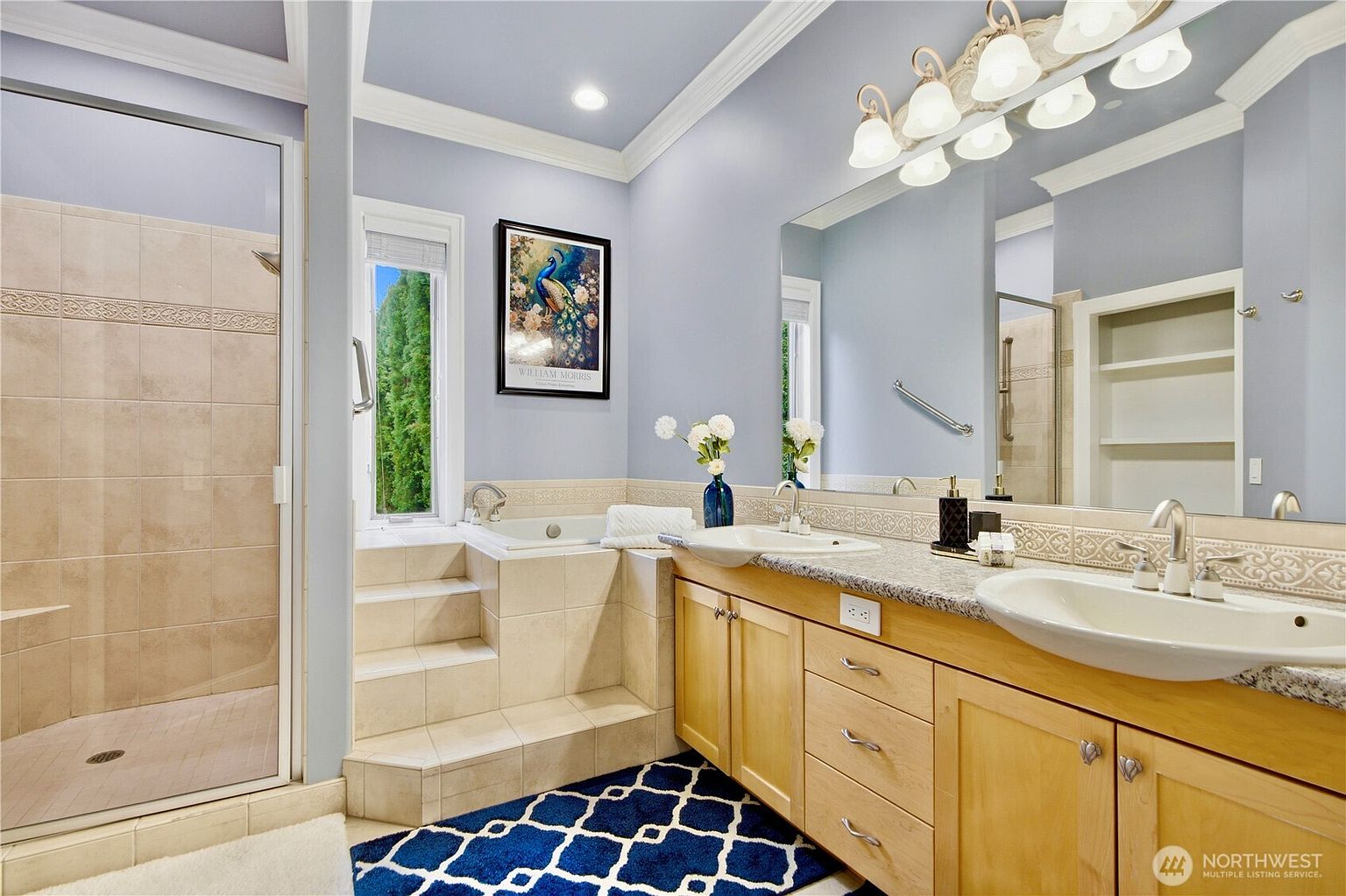 This primary bathroom features a dual-sink vanity with light wood cabinetry and a stone countertop, complemented by a large mirror and a multi-bulb light fixture. To the left, a tiled walk-in shower sits adjacent to a built-in soaking tub accessed by a set of steps. The room is painted in a soft blue, accented by a decorative tile border and a patterned blue and white rug on the floor.