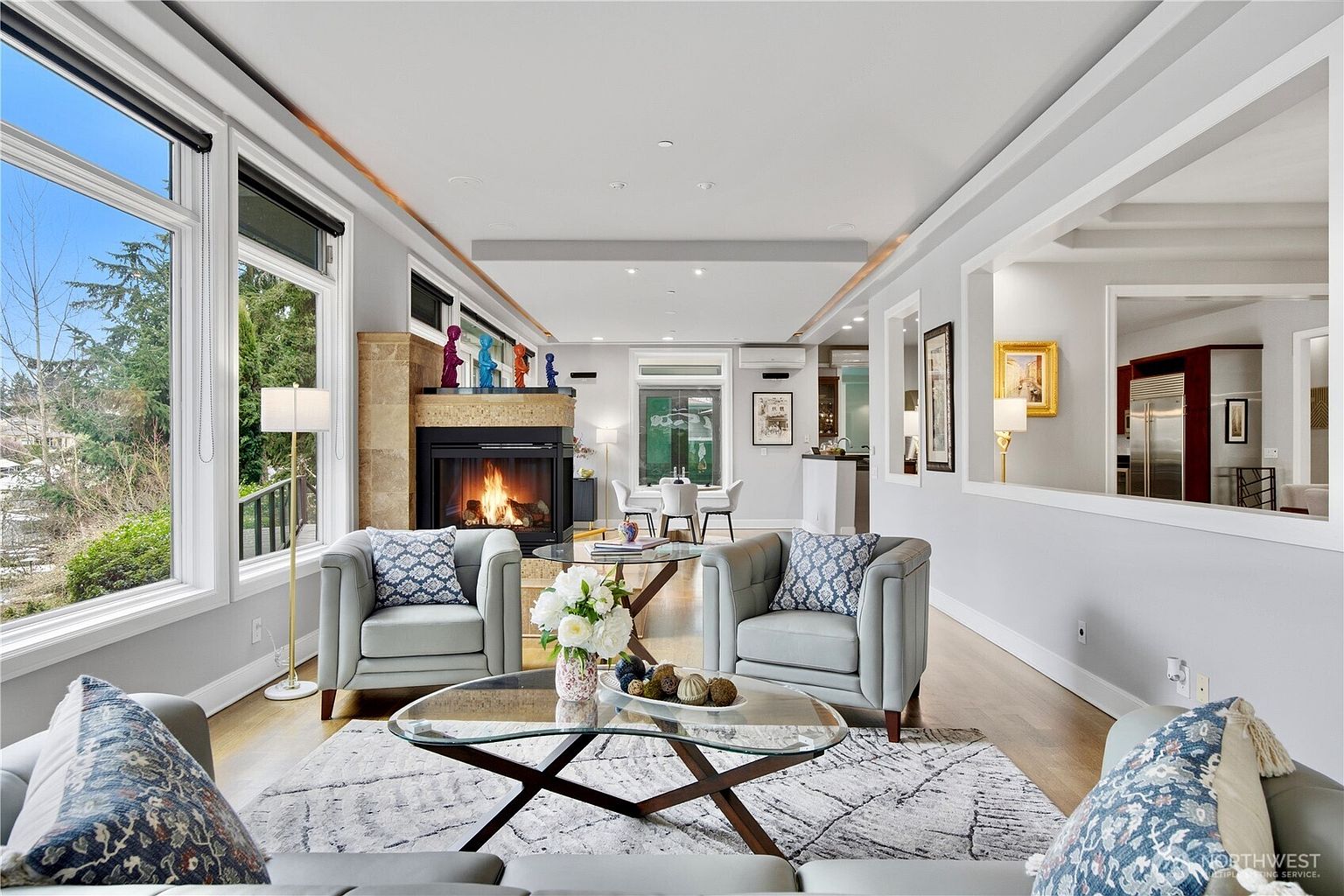 This bright and airy living room features a comfortable seating arrangement centered around a glass-topped coffee table and a stone-clad fireplace. Large windows along the left wall provide ample natural light and views of the outdoors, while a neutral color palette and patterned accent pillows create a sophisticated, inviting atmosphere. The open-concept layout flows seamlessly into a dining area and kitchen, highlighting the spacious and well-connected design of the home.