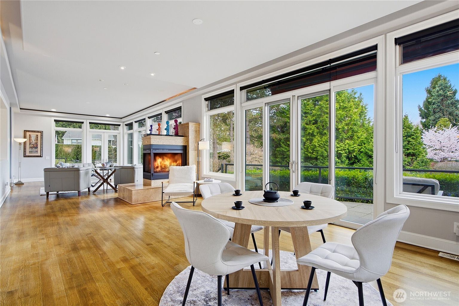 This bright and open-concept living and dining area features warm wood flooring and a central fireplace with a stone surround. Large glass sliding doors provide expansive views of the lush greenery outside, while a round dining table sits in the foreground on a circular rug. The space is designed with a neutral color palette and contemporary furnishings, creating an inviting and airy atmosphere.