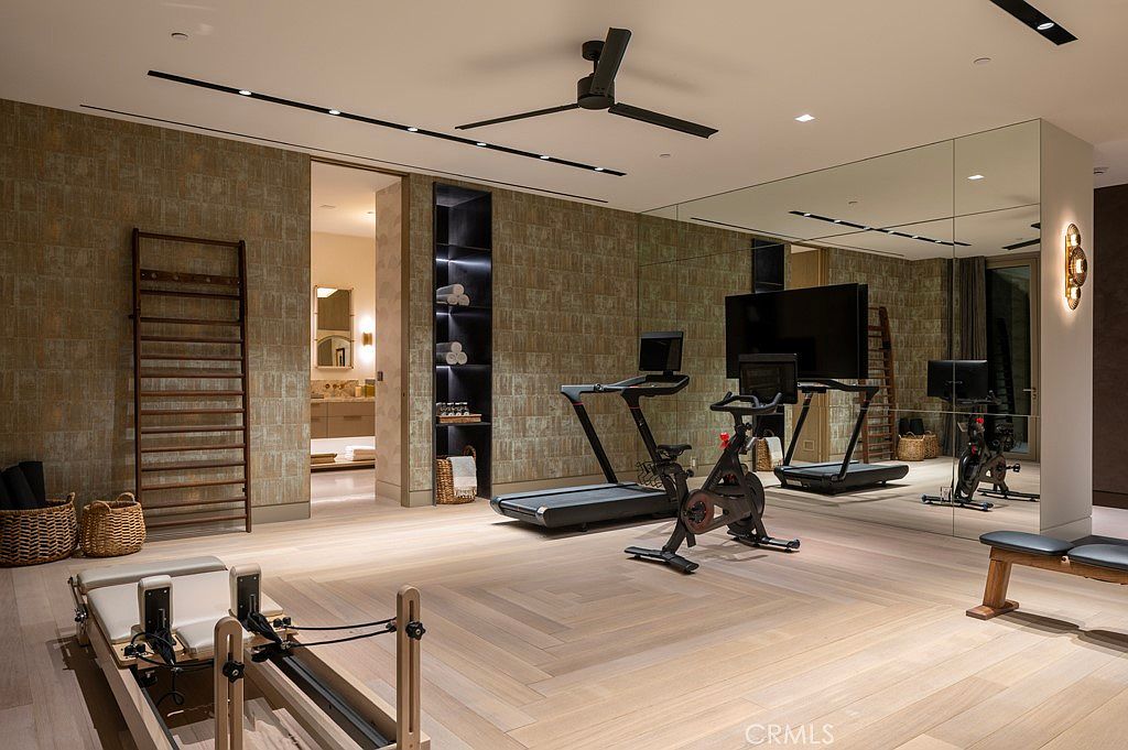 This is an interior shot of a home gym featuring a treadmill, stationary bike, and pilates reformer. The room has a modern aesthetic with wood flooring, textured wallpaper, and a large mirror wall that enhances the sense of space. A ceiling fan and built-in shelving add to the functionality and style of the room.