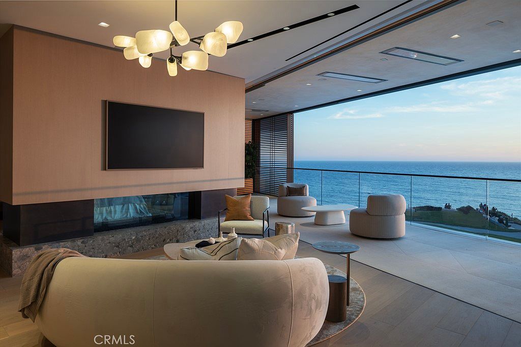 This is a luxurious living room with a stunning ocean view. The room features modern furniture, a sleek fireplace with a large television above, and a unique lighting fixture. The open floor plan and floor-to-ceiling windows create a bright and airy space, perfect for relaxation and entertaining.