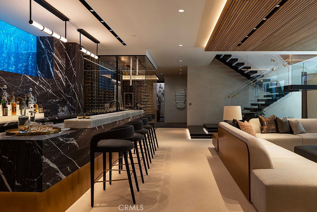 This interior shot showcases a luxurious living space featuring a modern bar area with marble countertops and stylish bar stools. The open floor plan seamlessly connects to a comfortable seating area with a plush sofa and decorative pillows, while a contemporary staircase with glass railings adds architectural interest. The overall impression is one of sophisticated elegance and comfortable living.