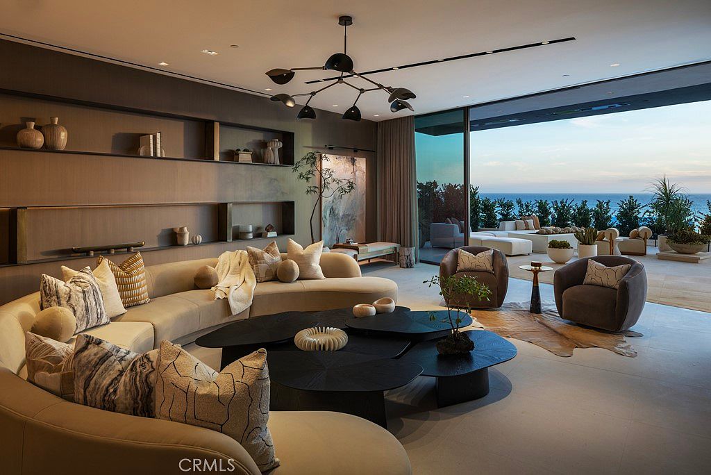 This is a luxurious living room featuring a curved sofa with decorative pillows, a unique multi-tiered coffee table, and stylish armchairs. The room has an open floor plan with large glass doors leading to an outdoor area with ocean views. The interior design is modern and sophisticated, creating a comfortable and elegant living space.