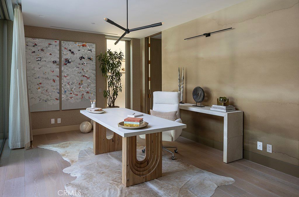 This is an interior shot of a modern office space featuring a white desk with wooden legs and a matching console table against a textured wall. The room is decorated with contemporary art and lighting fixtures, creating a sophisticated and functional workspace. A neutral-toned rug adds warmth to the hardwood flooring.