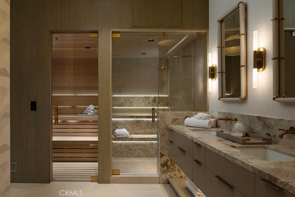 This is a luxurious primary bathroom featuring a sauna and a glass-enclosed shower with built-in seating. The vanity area has a marble countertop with dual sinks and modern lighting fixtures. The overall design is elegant and spa-like, creating a relaxing atmosphere.