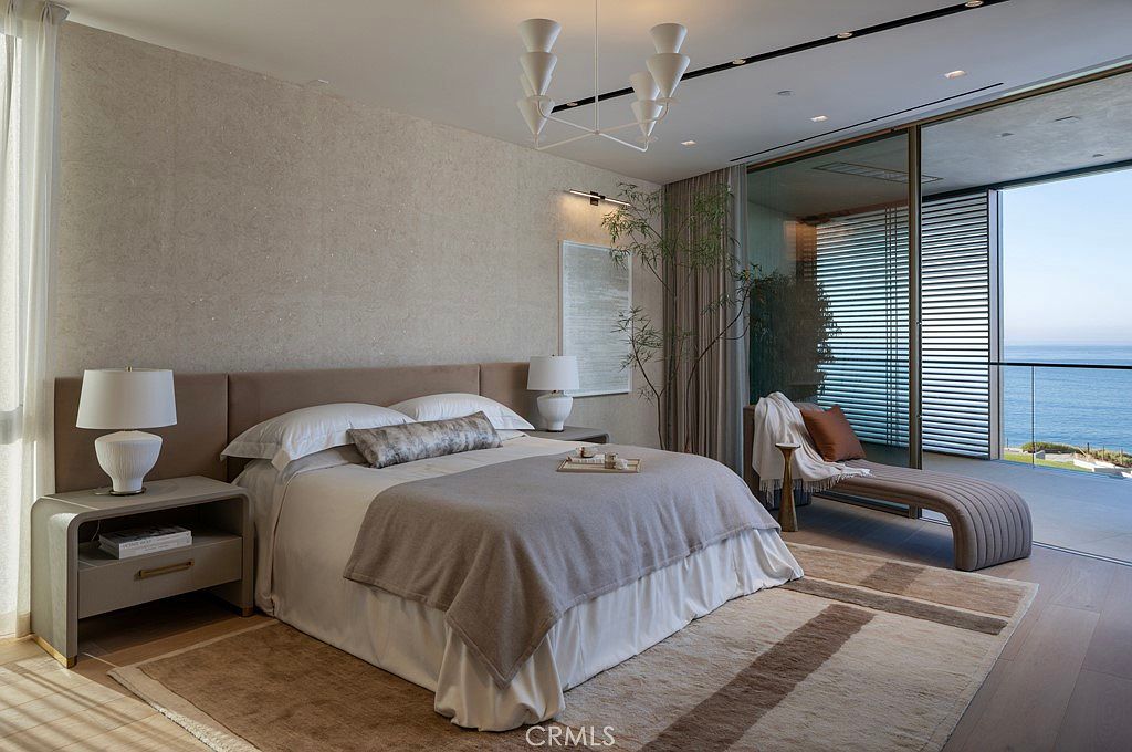 This is a luxurious primary bedroom featuring a king-size bed with a neutral-toned headboard and bedding, complemented by a stylish nightstand with a lamp. The room opens to a balcony with ocean views through large sliding glass doors, enhancing the sense of tranquility and high-end living. A modern chandelier hangs from the ceiling, adding a touch of elegance.