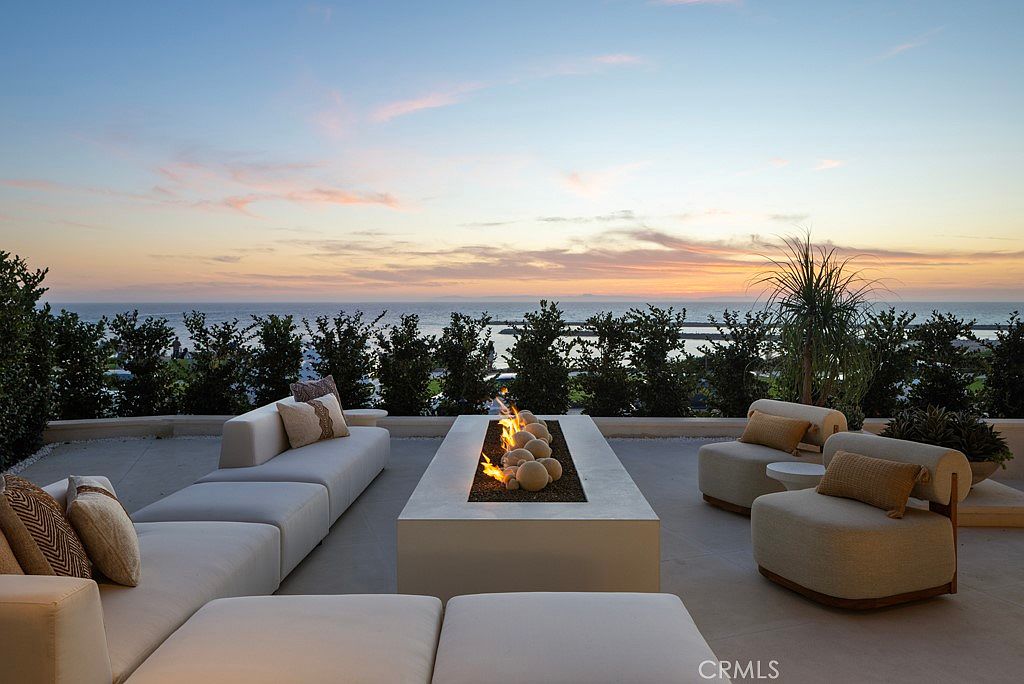 This image showcases a luxurious outdoor patio area at sunset, featuring a modern fire pit surrounded by comfortable lounge seating. The patio offers stunning ocean views framed by lush greenery, creating a serene and inviting atmosphere. The neutral color palette of the furniture complements the natural surroundings, enhancing the overall elegance of the space.