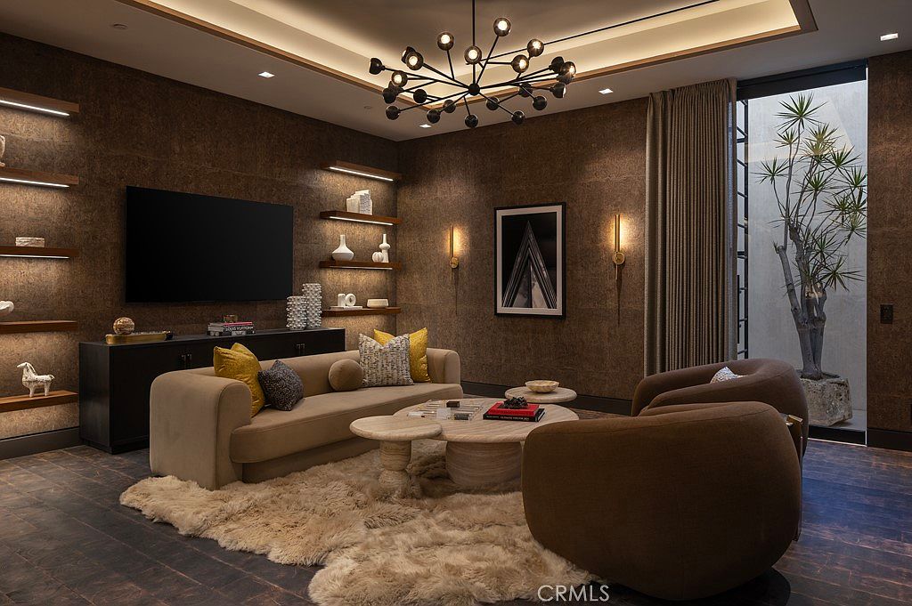 This is an interior shot of a living room featuring a modern design with warm, earthy tones. The room includes a beige sofa with decorative pillows, two round armchairs, and a unique multi-bulb chandelier. The walls are textured, and there are floating shelves with decorative items, creating a cozy and stylish atmosphere.