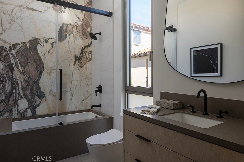 This is a well-appointed bathroom featuring a modern design with a bathtub and shower enclosed by a glass screen, showcasing marble-patterned walls. A sleek vanity with a dark countertop and light wood cabinetry complements the black fixtures, while a round mirror and a framed artwork add a touch of elegance. The room is brightened by natural light streaming through a window, creating a clean and inviting space.