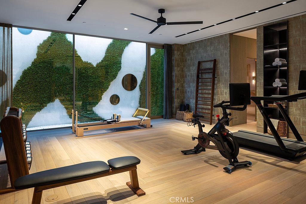 This is an interior shot of a modern home gym featuring a variety of exercise equipment including a stationary bike, treadmill, weight bench, and Pilates reformer. The room has light wood flooring, a large window with a unique green wall feature, and stylish wall coverings, creating a sophisticated and inviting workout space.