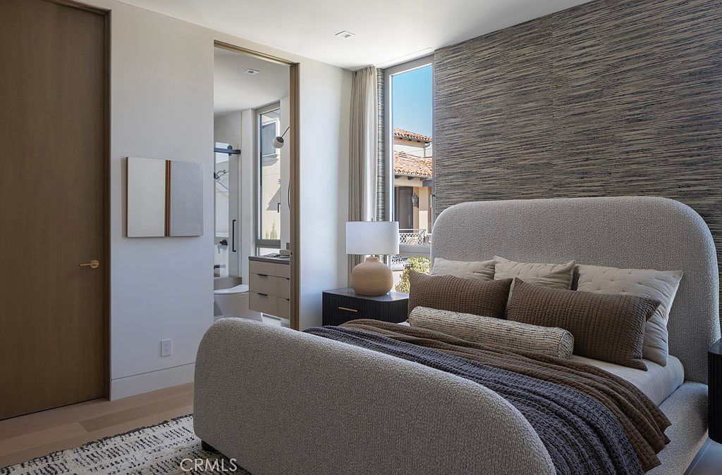 This is a primary bedroom featuring a large bed with a textured headboard and multiple pillows, creating a cozy and inviting atmosphere. The room has a modern aesthetic with a textured wall, a window providing natural light, and a glimpse into an adjacent bathroom. The color palette is neutral, contributing to a serene and sophisticated ambiance.
