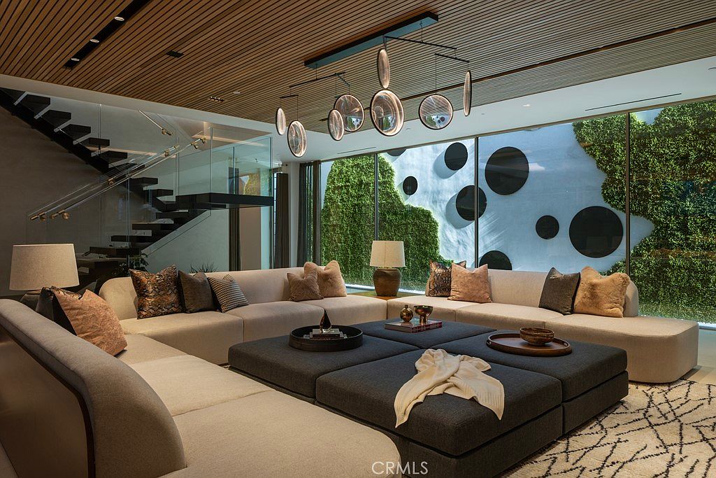 This is a luxurious living room featuring a large, sectional sofa with decorative pillows, a modular ottoman coffee table, and a modern chandelier. The room has a wall of windows overlooking a green space with artistic circular accents, and a staircase with glass railings is visible. The overall impression is one of contemporary elegance and comfort.