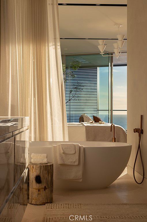 This is a luxurious primary bathroom featuring a freestanding bathtub with a view of the ocean. The room is decorated in neutral tones, with sheer curtains providing privacy while allowing natural light to filter in. A wooden stool sits next to the tub, adding a touch of rustic charm to the modern space.