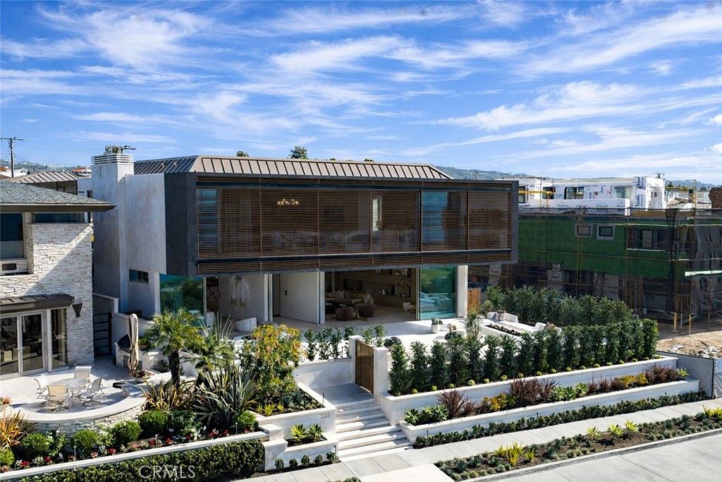 This is a front exterior view of a modern, two-story house with a unique architectural design. The house features a combination of concrete and wood elements, with large windows and a spacious outdoor patio area. The landscaping is well-maintained, with a variety of plants and shrubs adding to the property's curb appeal.
