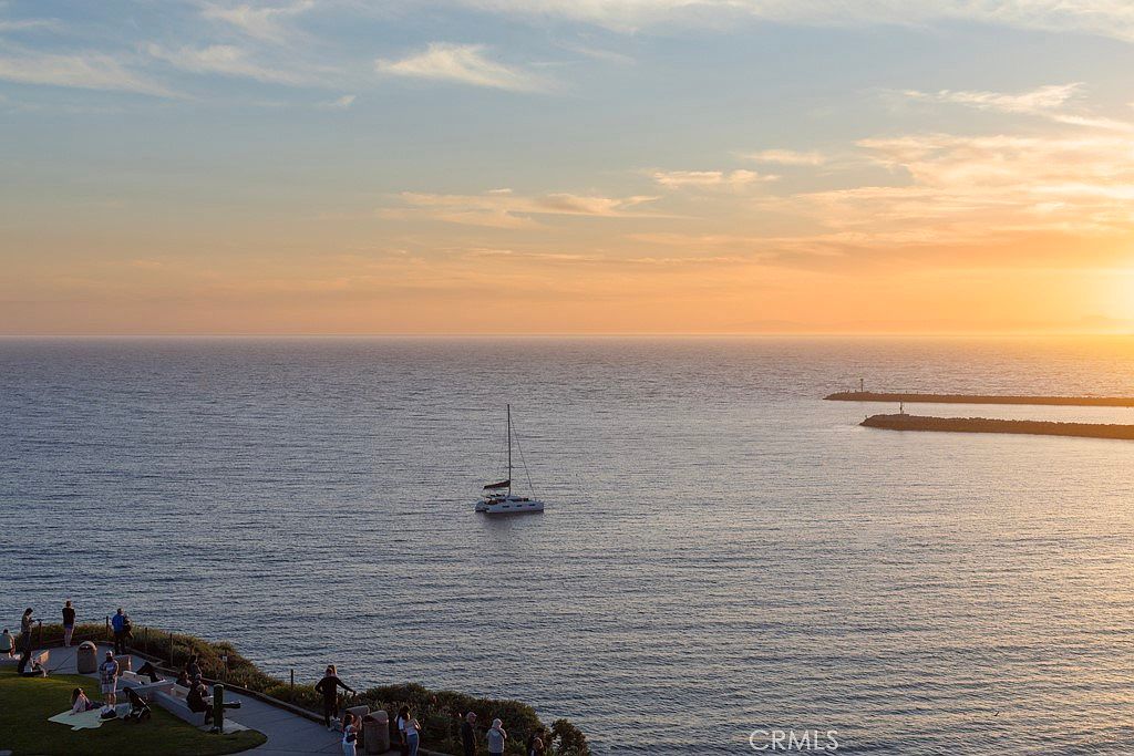 This image showcases a serene ocean view at sunset, featuring a sailboat gently sailing on the water. The scene is captured from an elevated perspective, overlooking a coastline with people enjoying the view. The warm hues of the sunset create a tranquil and inviting atmosphere, highlighting the beauty of the coastal landscape.