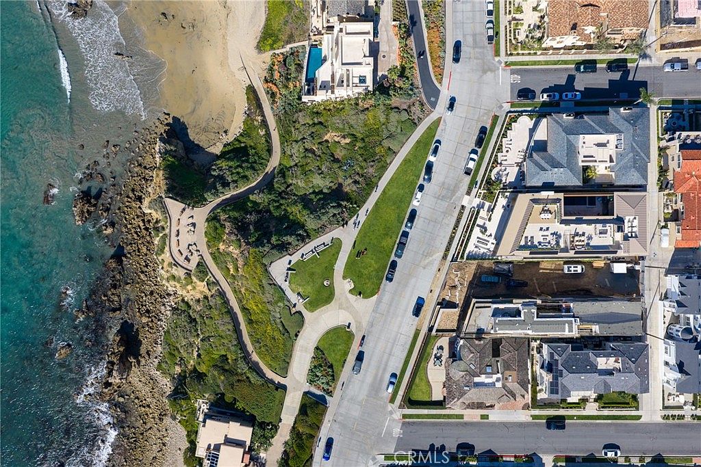 This aerial view showcases a coastal community with a blend of residential properties and natural landscapes. The scene features a rocky shoreline meeting the ocean, a sandy beach, and meticulously designed homes with varying architectural styles. A central road divides the properties from a green space with walking paths, creating a harmonious balance between urban living and natural beauty.