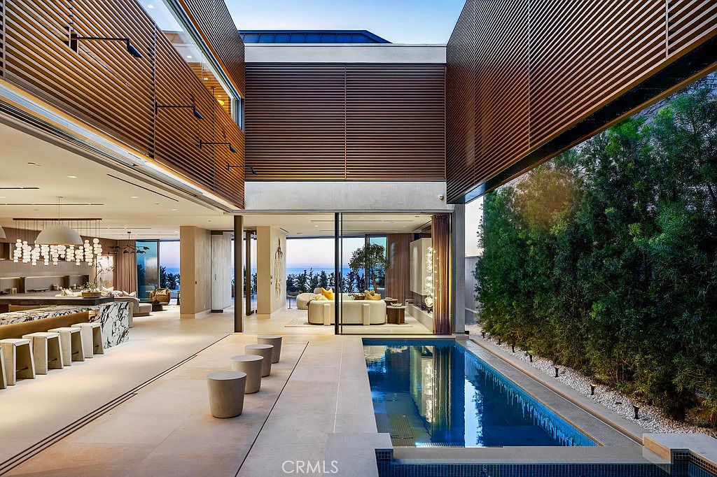 This image showcases a luxurious outdoor space featuring a sleek, narrow pool adjacent to a modern home. The pool's design integrates seamlessly with the home's architecture, which includes wooden slat details and large glass openings that blur the lines between indoor and outdoor living. The scene is framed by lush greenery, enhancing the sense of privacy and tranquility.