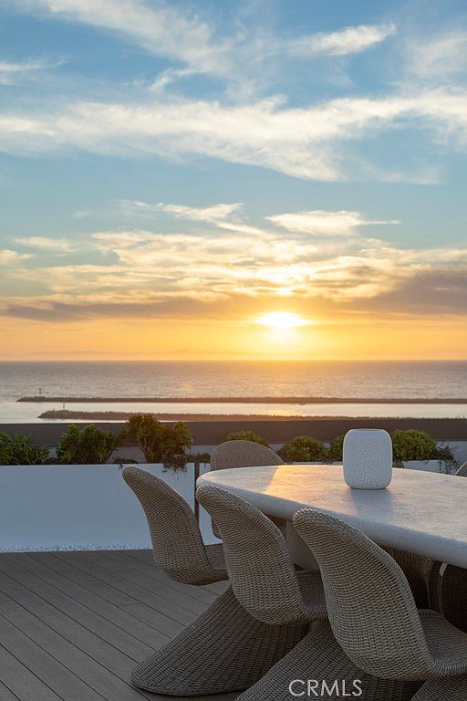 This image showcases a serene outdoor patio or balcony setting with a stunning ocean view at sunset. A modern outdoor dining set with woven chairs and a sleek table is prominently featured, suggesting a space for relaxation and entertainment. The warm hues of the sunset reflecting on the water create a luxurious and inviting atmosphere.