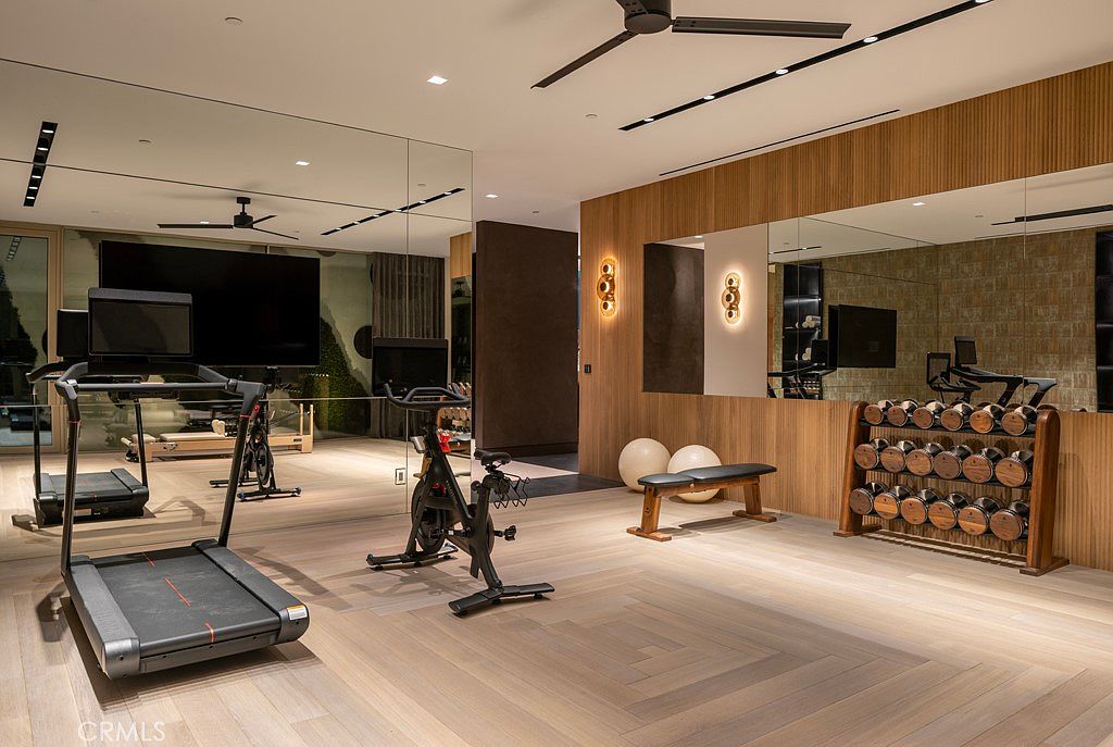 This is a well-appointed home gym featuring modern exercise equipment such as a treadmill and stationary bike. The room has a sleek design with wood paneling, mirrored walls, and a dumbbell rack. The flooring is light-colored wood, and the space is well-lit, creating an inviting atmosphere for workouts.