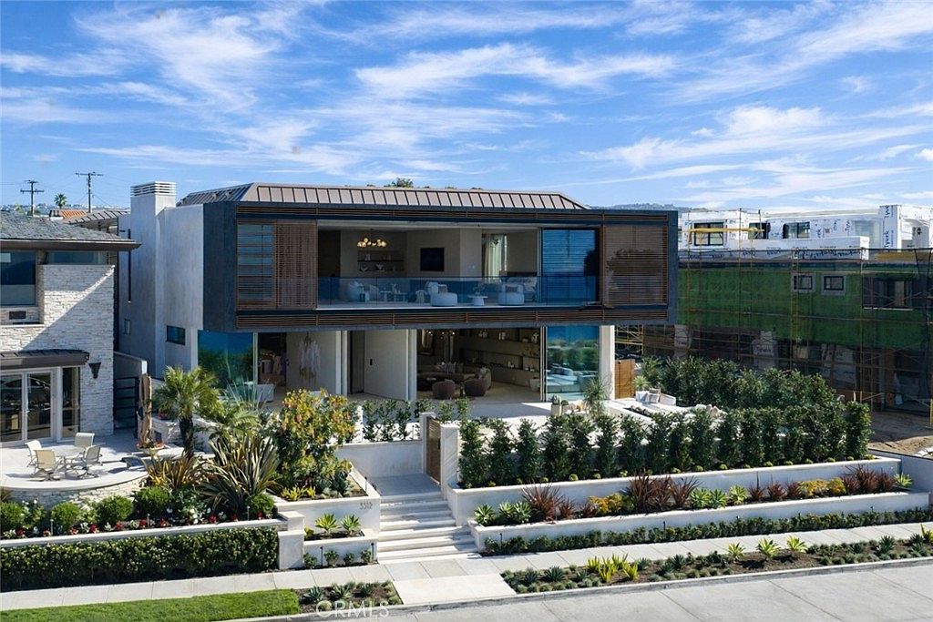 This is a front exterior view of a modern, two-story home. The house features a unique architectural design with a combination of concrete, wood, and glass elements. The landscaping includes tiered garden beds with lush greenery and a set of stairs leading to the entrance, creating an inviting curb appeal.