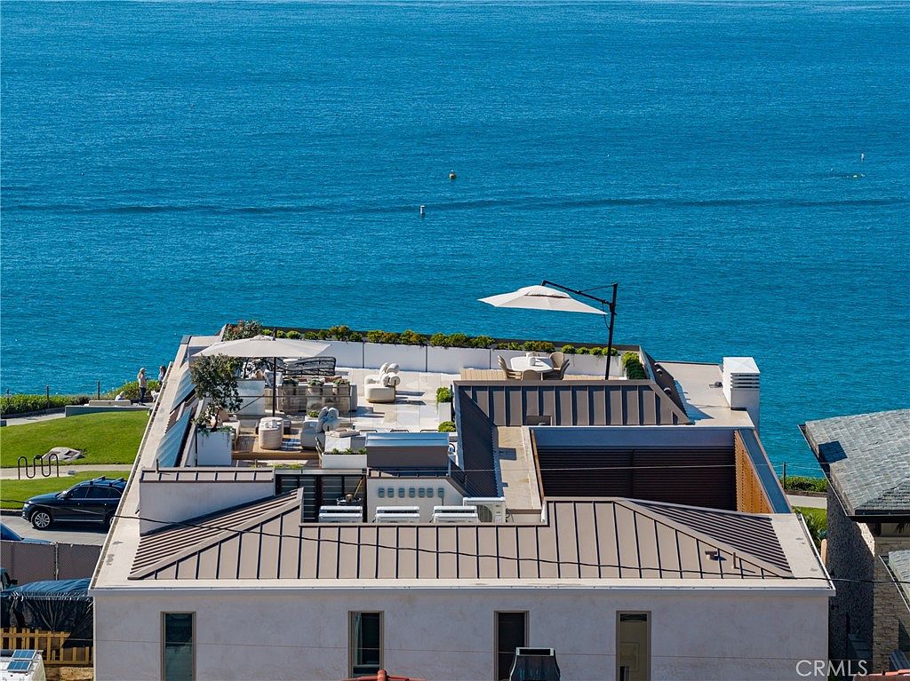 This aerial shot showcases a modern building with a rooftop deck featuring outdoor seating, umbrellas, and greenery, offering stunning ocean views. The building's architecture includes a mix of flat and gabled roofs, with a light-colored facade and dark trim, creating a sophisticated and inviting atmosphere. The surrounding landscape includes manicured lawns and neighboring structures, adding to the property's appeal.