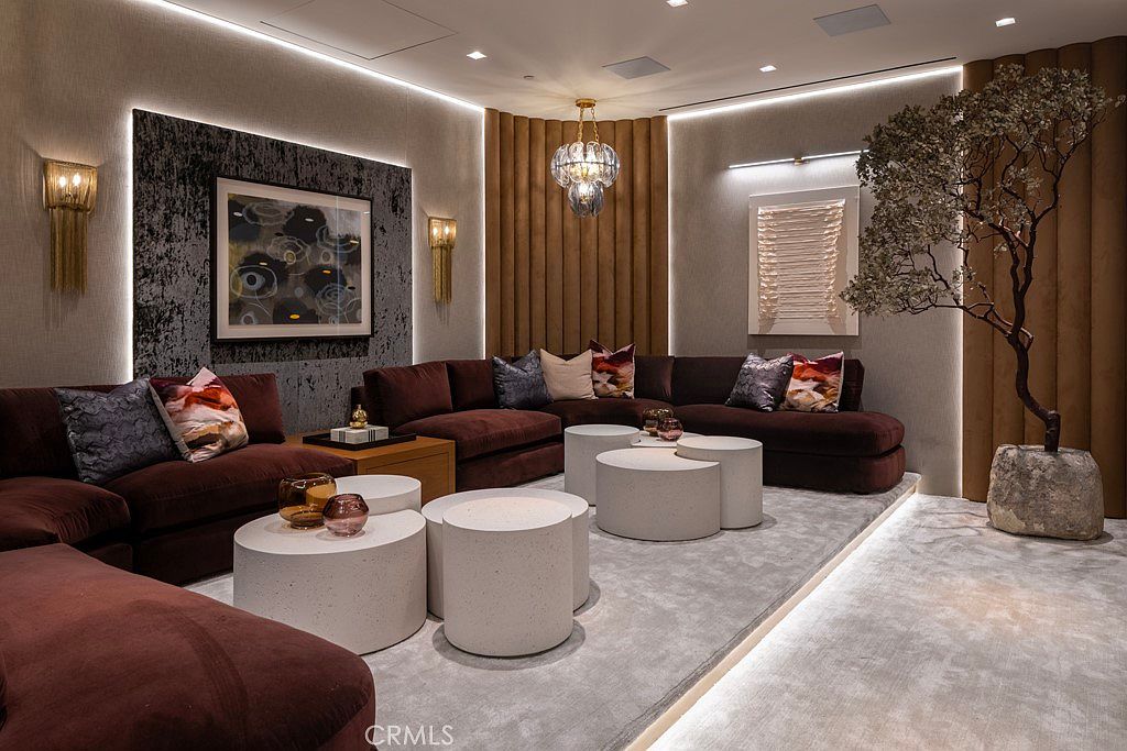 This is an interior shot of a luxurious living room featuring a large, sectional sofa in a deep burgundy color. The room is decorated with modern art, unique lighting fixtures, and several round, white coffee tables. A decorative tree adds a touch of nature to the space, creating a sophisticated and inviting atmosphere.