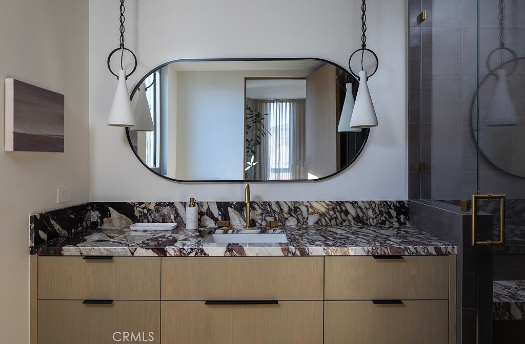 This is a primary bathroom featuring a modern vanity with a marble countertop and light wood cabinetry. An oval mirror with black trim hangs above the vanity, flanked by pendant lights. The bathroom also includes a glass-enclosed shower with marble accents, creating a luxurious and contemporary space.