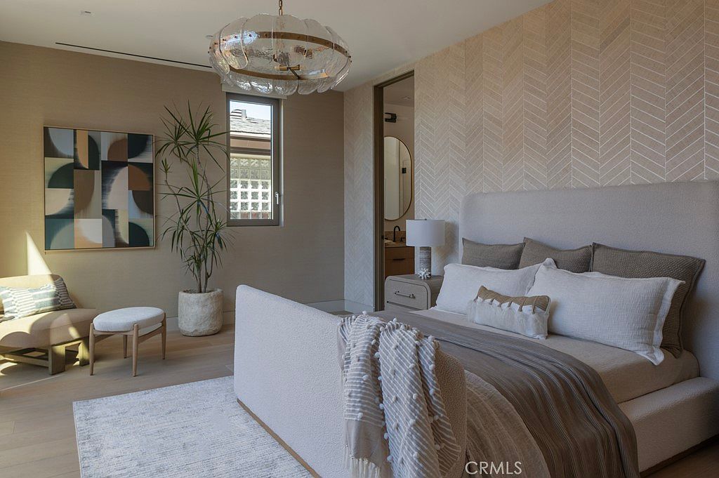 This is a serene primary bedroom featuring a neutral color palette with a textured accent wall. The bed is adorned with multiple pillows and a cozy throw, complemented by a stylish chandelier and an abstract art piece. A potted plant adds a touch of nature, enhancing the room's tranquil atmosphere.