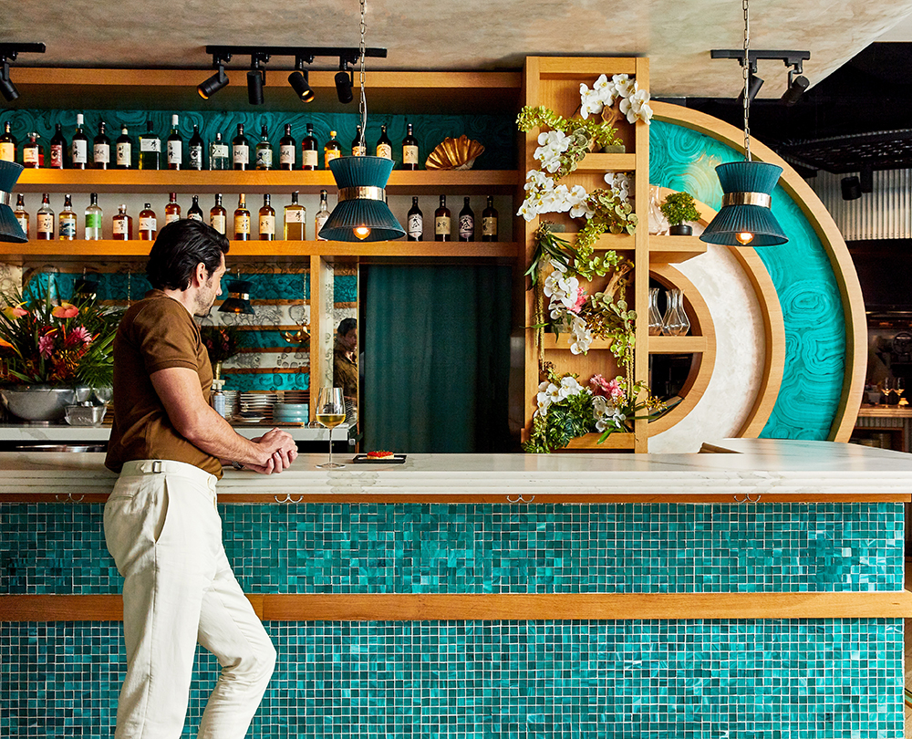 This interior shot showcases a stylish dining area with a unique bar design featuring teal mosaic tiles and wooden accents. A man stands at the bar, adding a sense of scale and activity to the scene. The lighting is warm and inviting, highlighting the decorative elements such as the floral arrangements and the curated selection of bottles on the shelves.