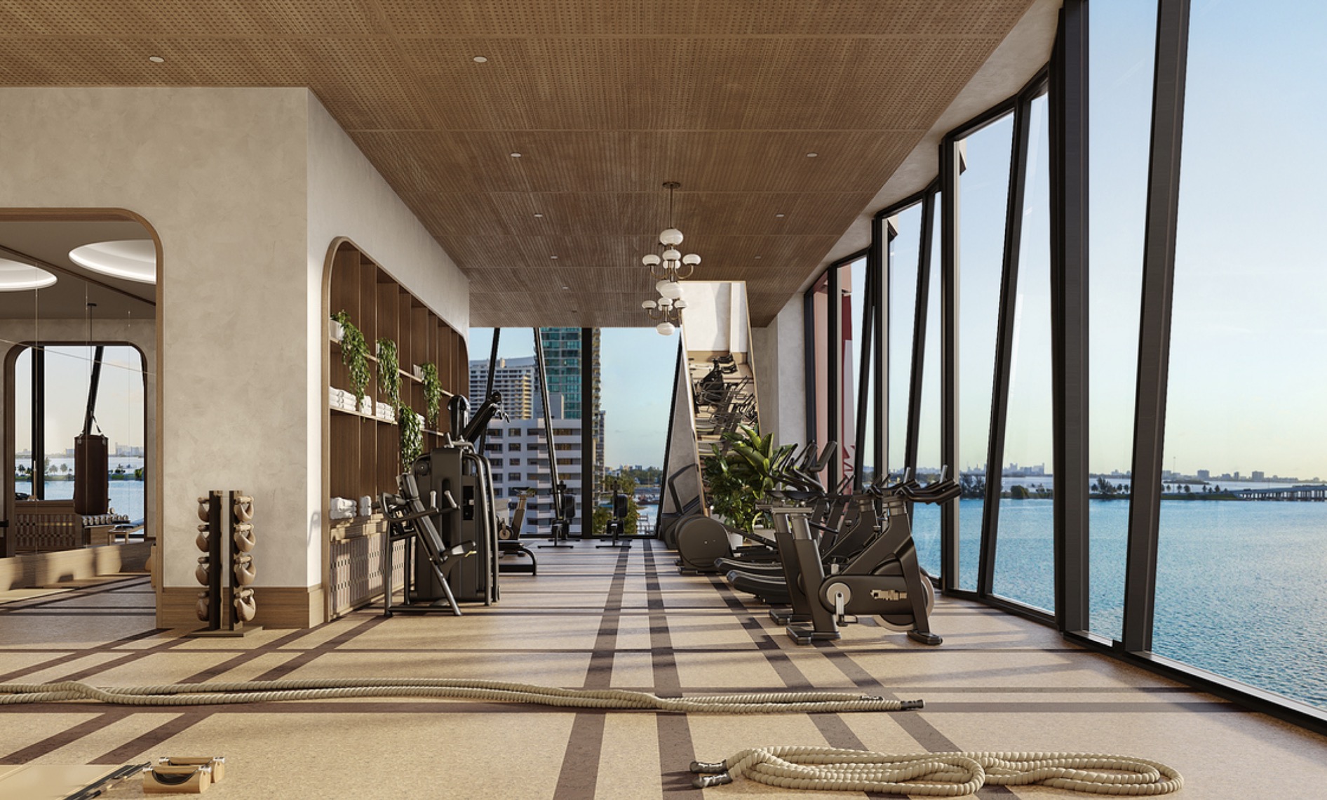 This is a modern home gym featuring state-of-the-art exercise equipment and floor-to-ceiling windows offering stunning water views. The space is well-lit with a wood-paneled ceiling and includes architectural details like arched doorways and built-in shelving with greenery. The overall impression is one of luxury and wellness.
