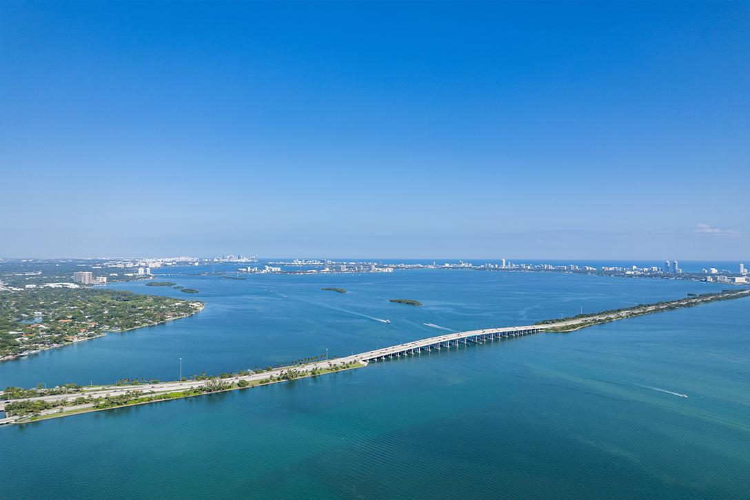 This aerial shot showcases a stunning coastal landscape with a long bridge stretching across the turquoise water, connecting lush green islands. The cityscape in the distance adds a touch of urban sophistication to the serene natural setting. The clear blue sky enhances the overall sense of openness and tranquility, making it an appealing location.