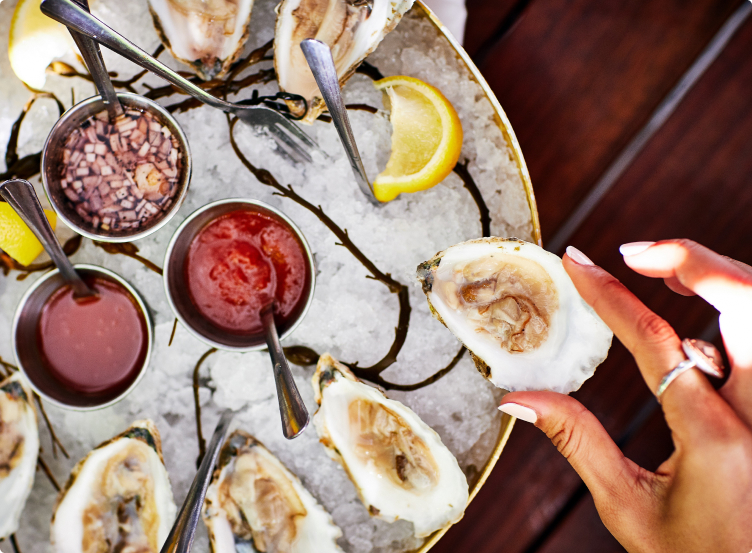This image showcases a luxurious food presentation, likely intended to highlight a high-end dining experience. A selection of oysters on ice is arranged artfully on a platter, accompanied by various sauces and lemon wedges. A hand delicately holds one of the oysters, suggesting freshness and quality, creating an inviting and upscale impression.