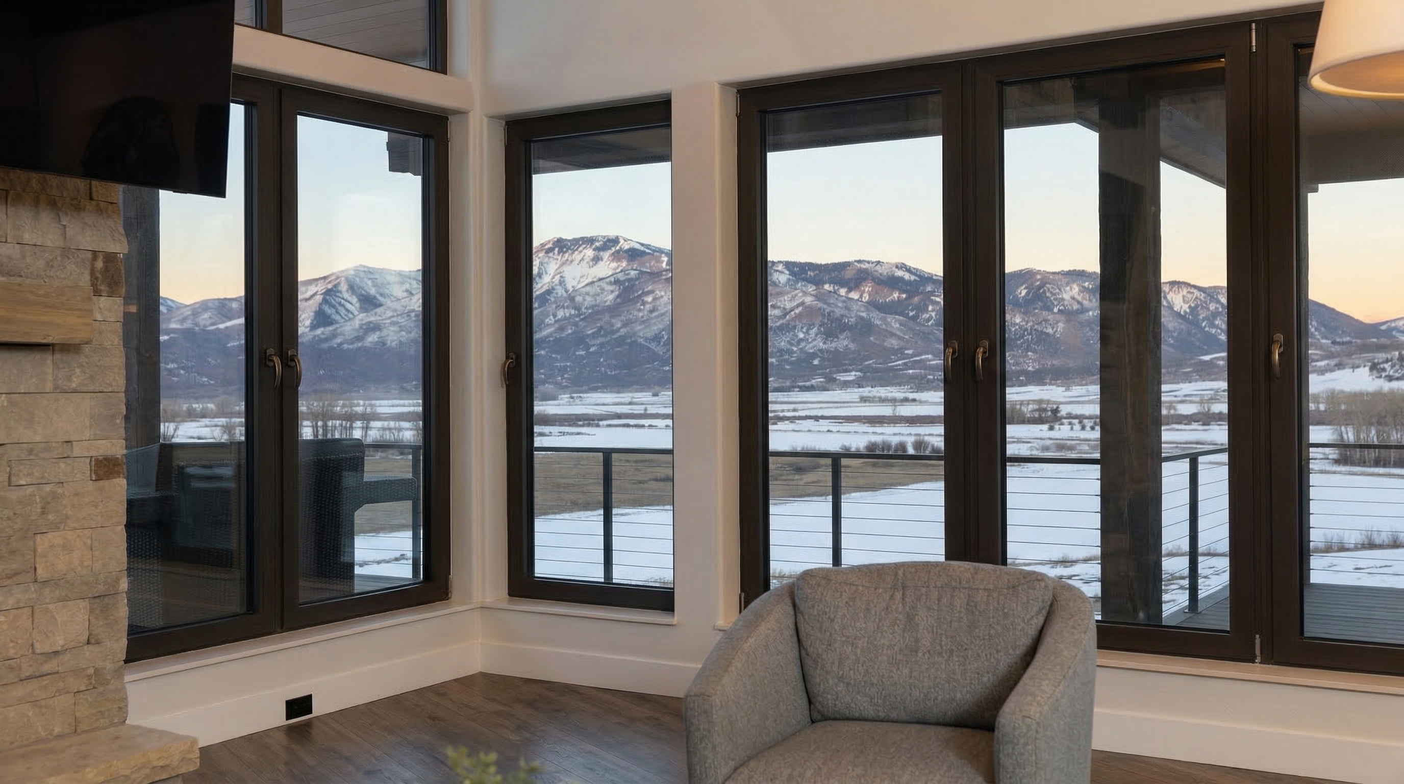 This living room features a striking stone fireplace adjacent to floor-to-ceiling windows that provide expansive, panoramic views of snow-capped mountains and a winter landscape. The space is highlighted by a cozy grey accent chair, sleek dark-framed windows, and warm wooden flooring, creating a sophisticated and serene atmosphere. It is an ideal space for relaxing and taking in the spectacular scenery.