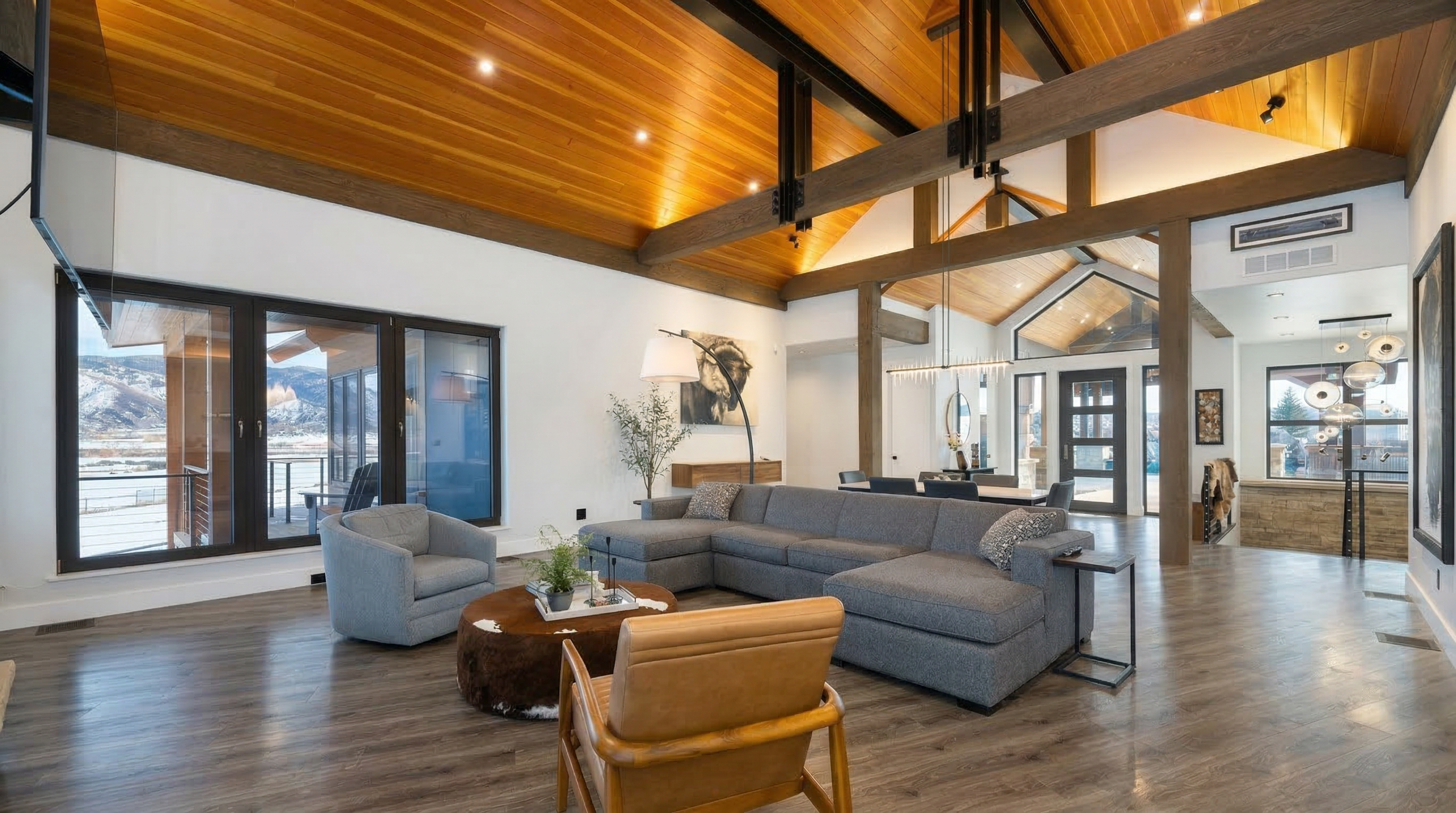 This spacious, open-concept living room showcases a striking vaulted ceiling with warm wood paneling and exposed timber beams. The area is anchored by a large, comfortable gray sectional, a circular cowhide-textured ottoman, and a leather armchair, all illuminated by recessed lighting and a sleek arched floor lamp. Large glass sliding doors frame a scenic view of snow-covered mountains, emphasizing the home's bright, modern mountain-lodge aesthetic.