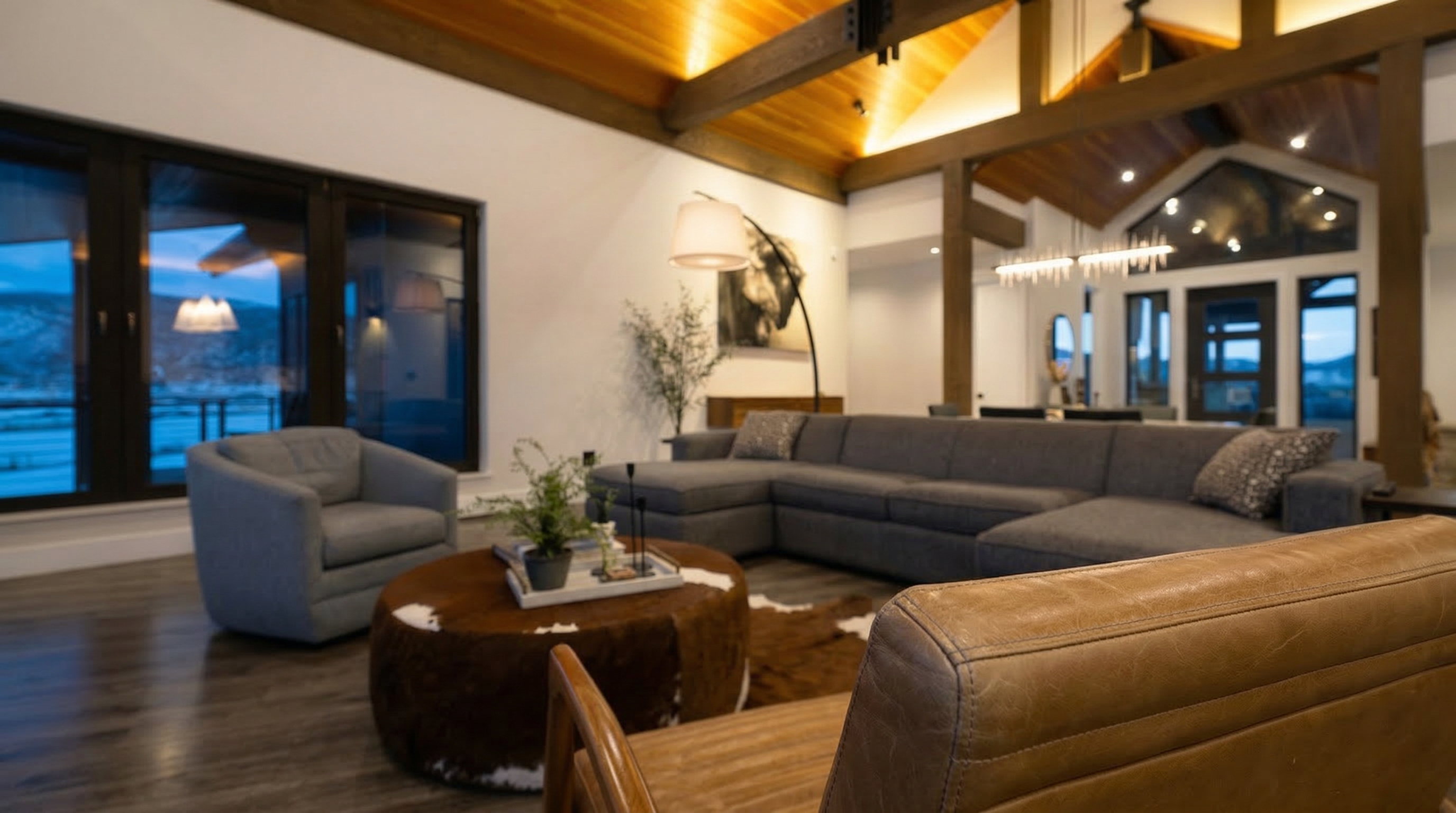 This spacious and inviting living room features a vaulted ceiling with exposed timber beams and warm recessed lighting, creating a cozy yet sophisticated atmosphere. The open floor plan showcases a large grey sectional sofa, a round cowhide-style coffee table, and a modern floor lamp, all centered around a contemporary aesthetic. Expansive windows provide ample natural light and a scenic view, enhancing the room's high-end, comfortable rustic charm.