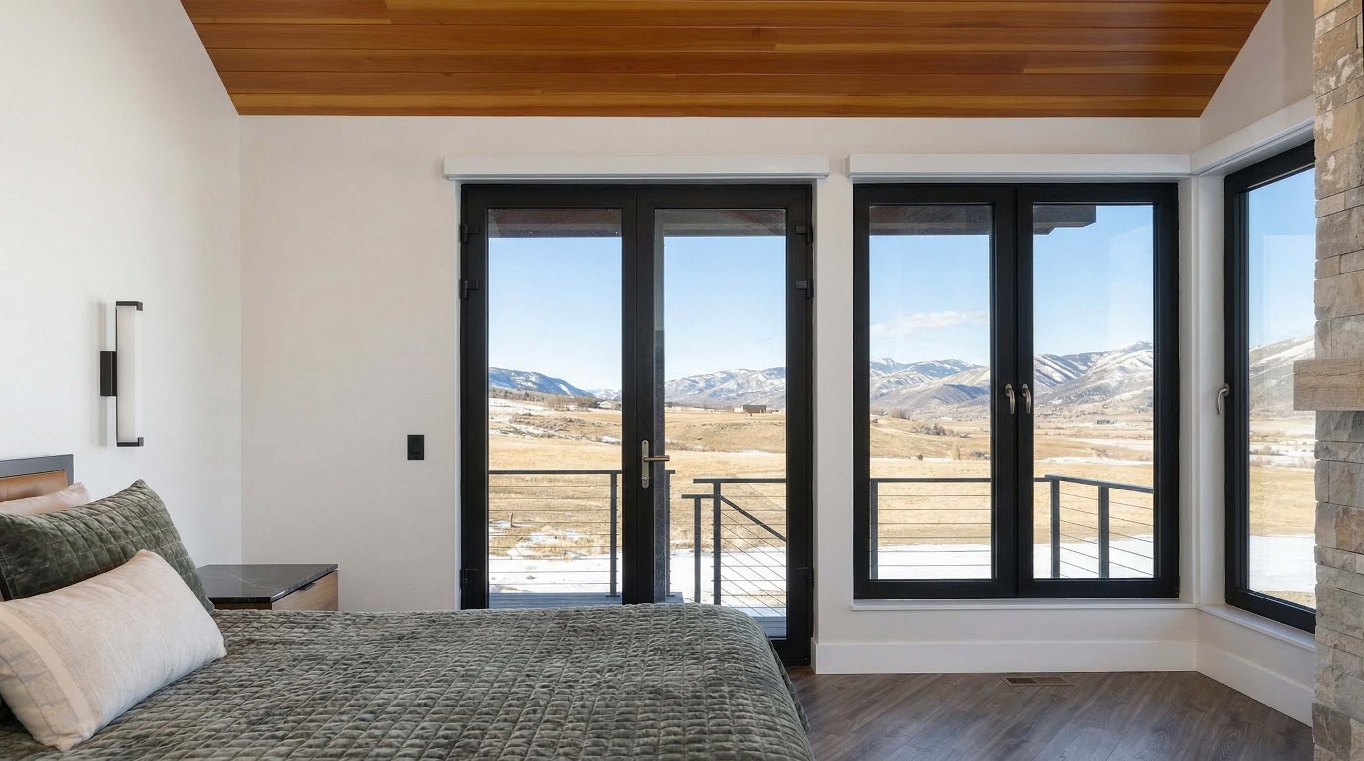 This modern primary bedroom features a striking vaulted ceiling with warm wood tongue-and-groove paneling and expansive floor-to-ceiling black-trimmed windows that perfectly frame a stunning panoramic view of snow-capped mountains. The interior design blends rustic warmth with a contemporary aesthetic, highlighted by a textured olive-green quilt and clean white walls. The space offers a seamless connection to the outdoors, creating a serene and luxurious retreat.