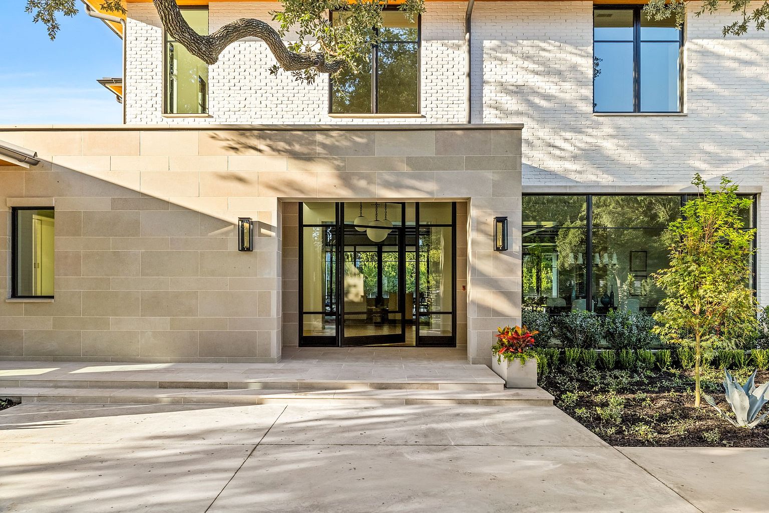 This is a front exterior shot of a modern home, emphasizing the entryway. The facade features a combination of light-colored brick and stone, with large windows and a striking black-framed glass door. The landscaping is well-maintained, adding to the property's curb appeal and creating an inviting atmosphere.