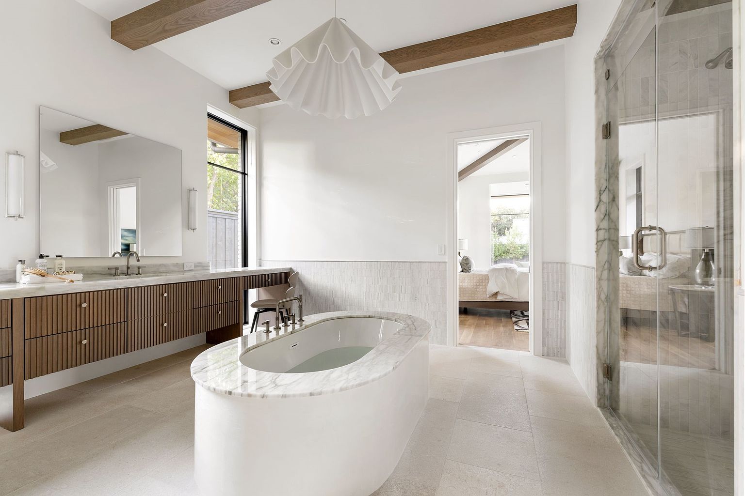 This is a bright and luxurious primary bathroom featuring a freestanding oval bathtub with a marble surround, a long wooden vanity with a marble countertop, and a glass-enclosed shower. The room is decorated in a neutral color palette with wooden beams on the ceiling, creating a spa-like atmosphere. The perspective is from the doorway, showcasing the entire space.