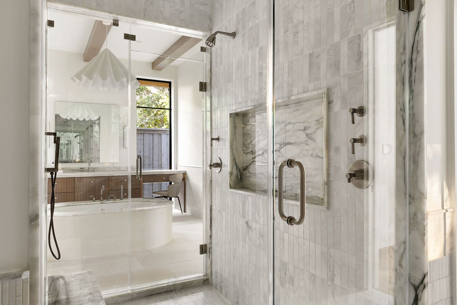 This is a luxurious primary bathroom featuring a glass-enclosed shower with marble tile walls and a built-in niche. The bathroom also includes a freestanding oval bathtub, a vanity with wood cabinetry, and a large mirror. The overall design is elegant and modern, creating a spa-like atmosphere.