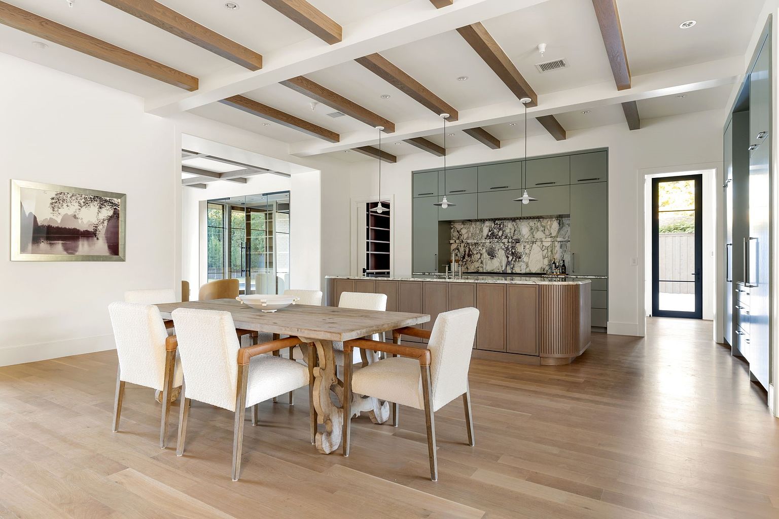 This interior shot showcases a dining room adjacent to a modern kitchen. The dining area features a rustic wooden table surrounded by upholstered chairs, complemented by exposed wooden beams on the ceiling. The kitchen boasts sleek cabinetry and a marble backsplash, creating a sophisticated and inviting atmosphere.