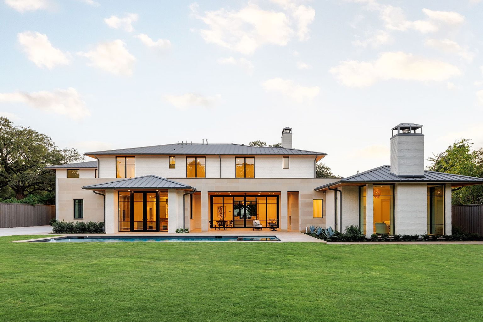 This is a rear view of a modern, luxurious home featuring a pristine green lawn, a swimming pool, and a light-colored brick exterior. Large glass doors and windows offer ample natural light and create a seamless transition between indoor and outdoor living spaces. The architecture is contemporary with clean lines and a sophisticated aesthetic.