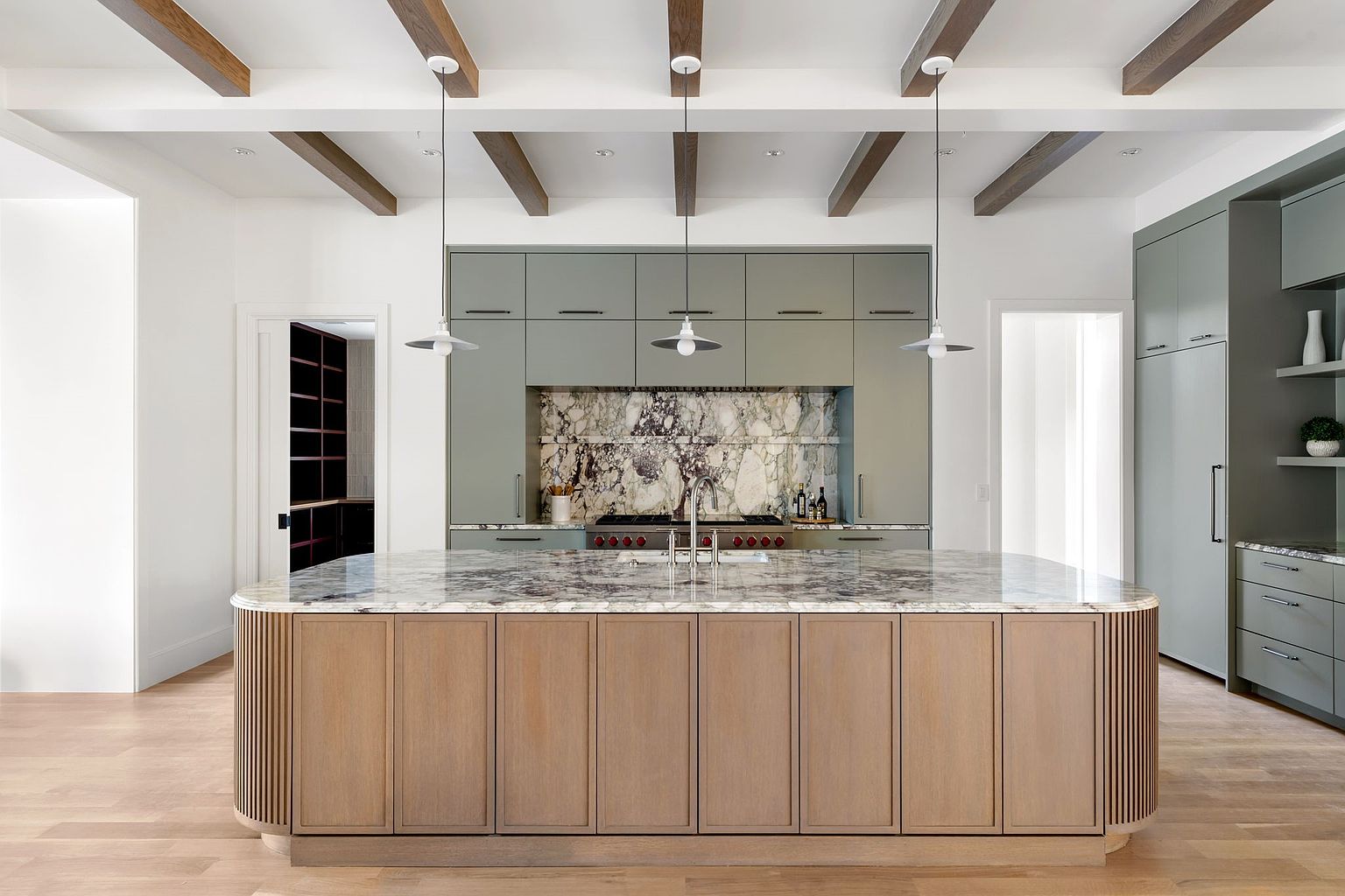 This is a well-lit, modern kitchen featuring a large island with a marble countertop and wooden cabinetry. The kitchen also includes sage green cabinets, a marble backsplash, and wooden beams on the ceiling. The overall impression is one of luxury and sophistication.