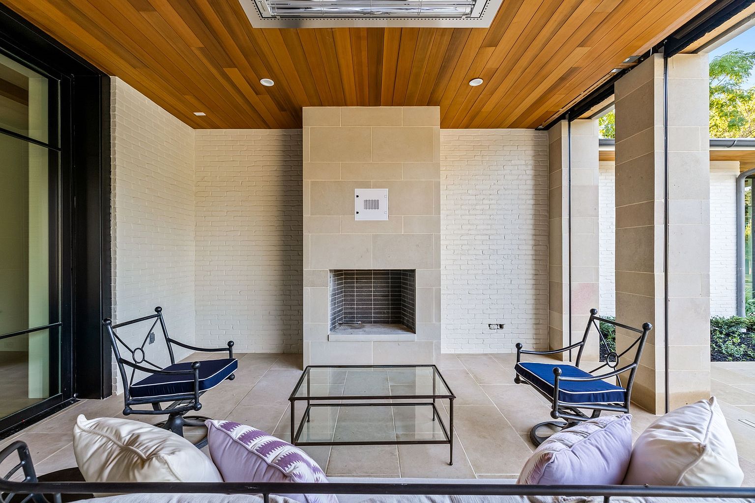This image showcases a covered patio area with a modern design. The space features a stone fireplace, comfortable seating with plush cushions, and a glass-topped coffee table. The ceiling is finished with wood planks, and the overall impression is one of luxury and relaxation, perfect for outdoor entertaining.