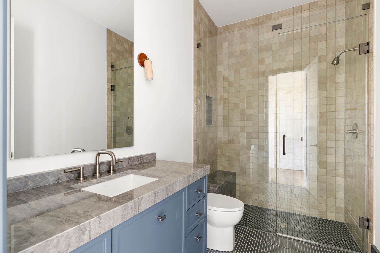 This is a primary bathroom featuring a vanity with a gray marble countertop and blue cabinetry. A large mirror hangs above the sink, and a modern sconce provides lighting. The shower is enclosed with glass and tiled in neutral tones, with a built-in bench and a contrasting dark gray tiled floor, creating a spa-like atmosphere.