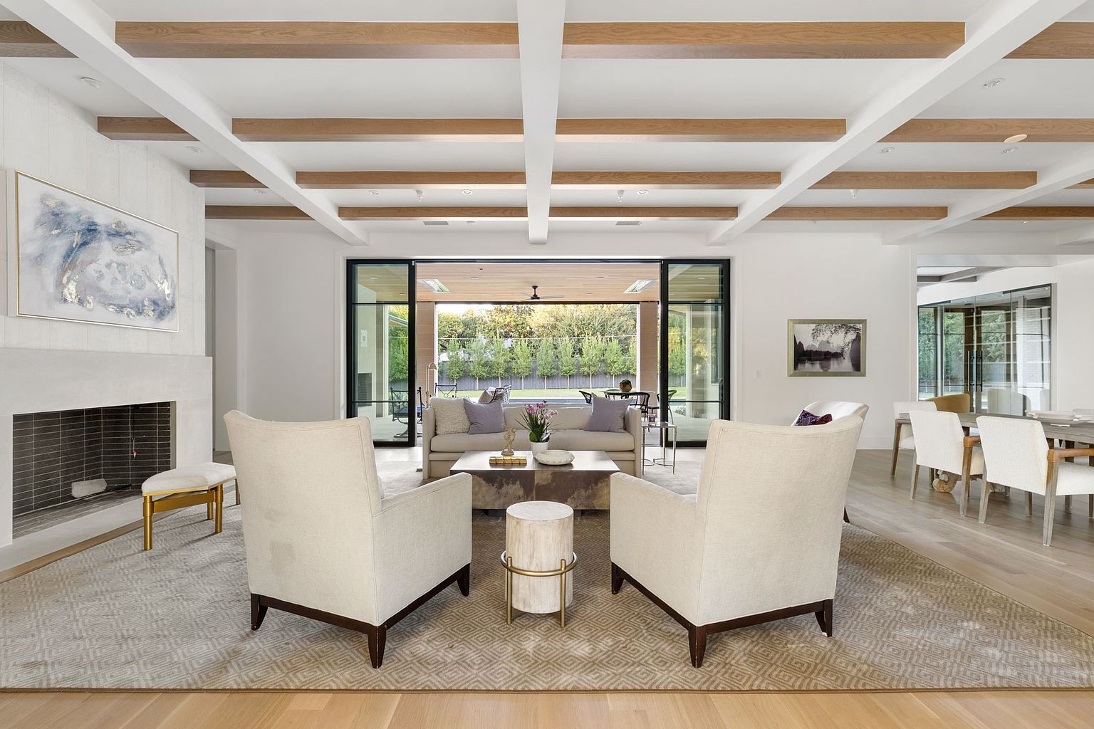 This is an interior shot of a living room featuring a neutral color palette with white walls and light wood flooring. The room is furnished with two light-colored armchairs, a sofa, and a coffee table, arranged around a fireplace. Exposed wooden beams adorn the ceiling, adding a touch of rustic charm to the modern design, and large sliding glass doors open to an outdoor space, providing ample natural light and a seamless indoor-outdoor transition.