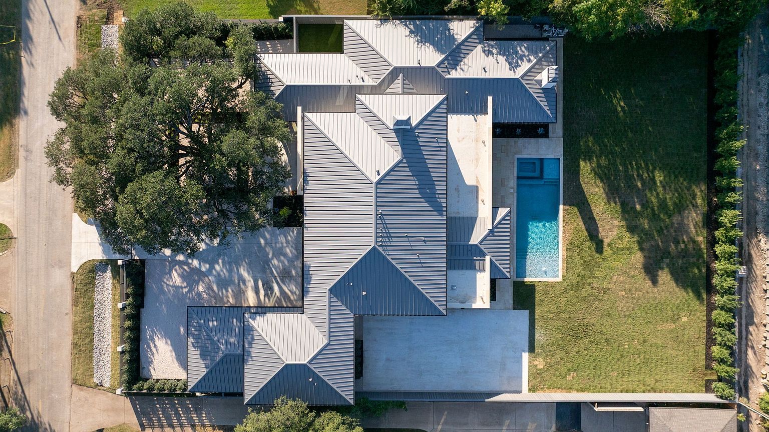 This aerial shot showcases a modern home with a complex, multi-gabled roof design in varying shades of gray. A rectangular swimming pool is visible adjacent to the house, surrounded by a well-manicured lawn. The property also features a spacious driveway and mature trees, adding to its curb appeal and privacy.