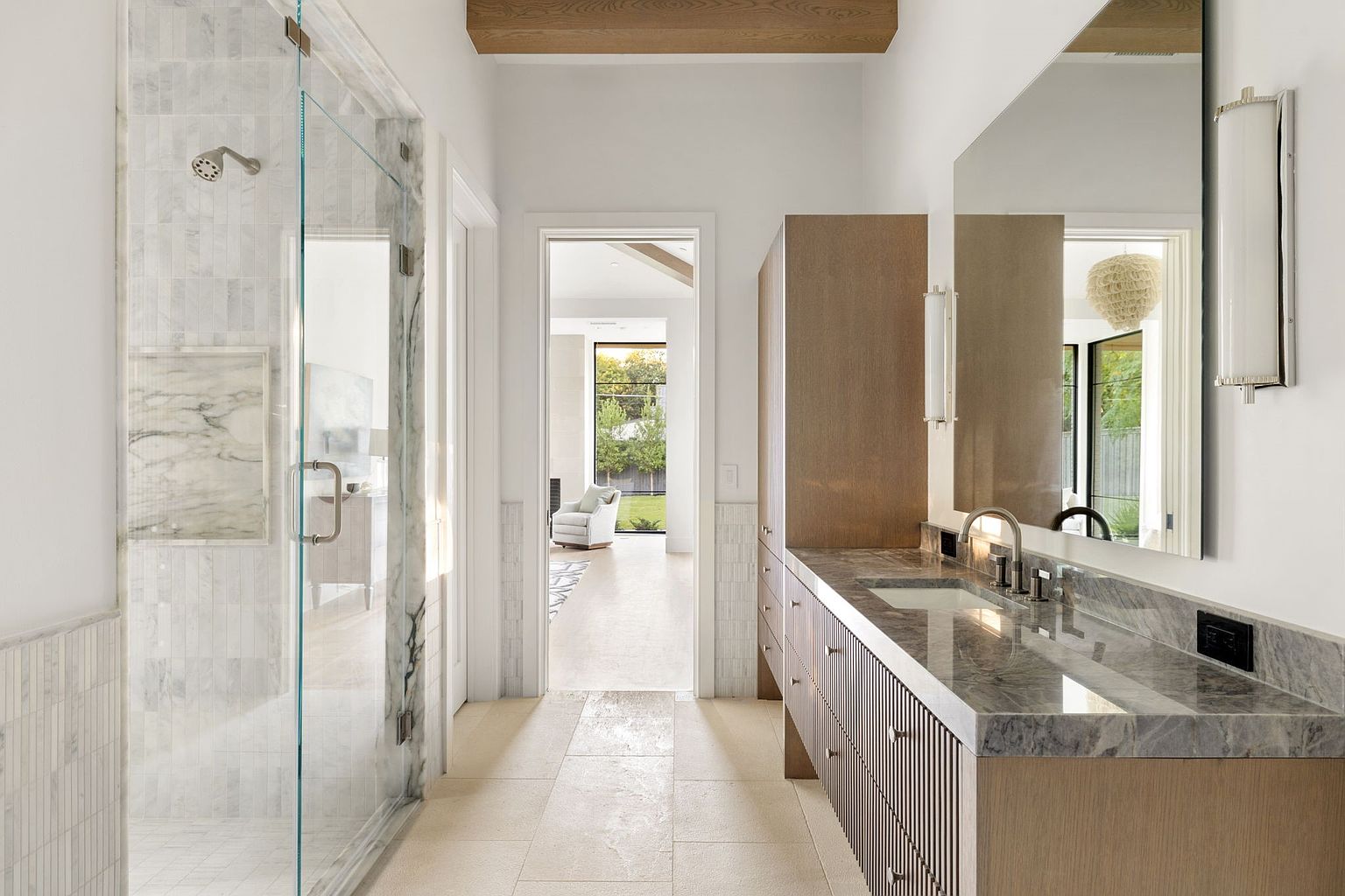 This is a bright and modern primary bathroom featuring a glass-enclosed shower with marble tiling, a long vanity with a gray countertop and wooden cabinets, and a large mirror reflecting the adjacent room. The neutral color palette and clean lines create a spa-like atmosphere, perfect for relaxation and luxury. The hallway leads to another room with a view of the backyard.